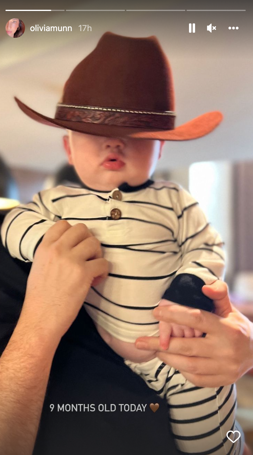Nine-month old Malcolm wears a striped pajama set and wears an oversized cowboy hat that covers his eyes. He's being held by an adult that's not in frame. Olivia Munn and John Mulaney's baby.